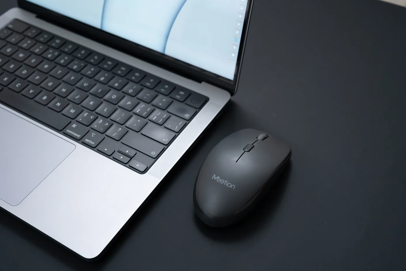 Meetion BTM100 mouse next to a laptop on a dark desk